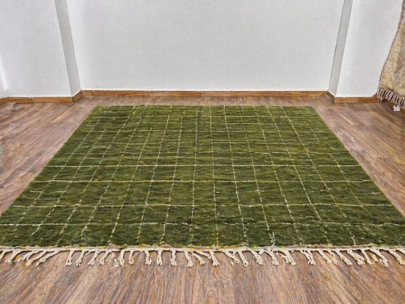 May include: A rectangular green rug with a grid pattern and cream-colored fringe. The rug is made of a textured material and is placed on a wooden floor. The grid pattern is created by white lines.