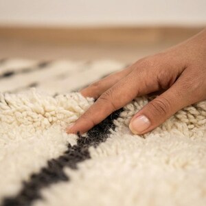 May include: A thick, cream-colored shag rug featuring bold, dark charcoal-gray horizontal stripes. The rug has a deep, plush pile texture with long, soft fibers that create a dense and cozy surface appearance across the entire woven textile.