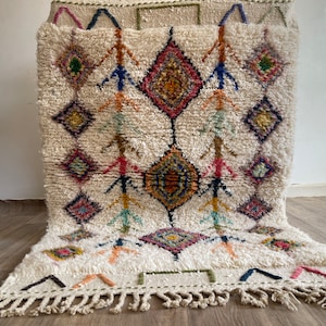 May include: A white Moroccan rug with a geometric pattern in shades of pink, blue, green, and yellow. The rug has a fringe border and is made of wool.