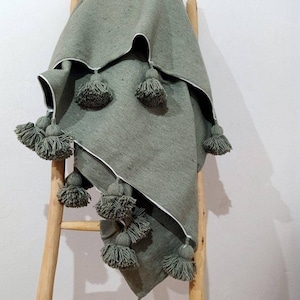 Pode incluir: Uma manta de arremesso verde-sálvia com uma borda branca e grandes pompons ao redor das bordas. A manta está drapeada sobre uma escada de madeira.