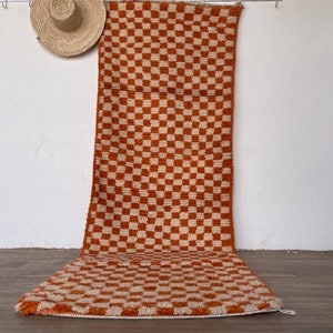 May include: A long, rectangular rug with a checkered pattern in shades of orange and white. The rug is made of a soft, fluffy material and has a fringe border.