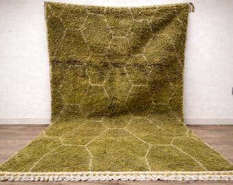 May include: A large, olive green rug with a geometric pattern of interconnected hexagons outlined in white. The rug has a shaggy texture and a white and yellow fringe along the bottom edge. It is displayed against a white wall.