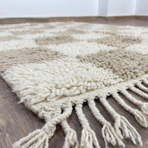 May include: A beige and cream-colored rug with a geometric pattern. The rug has a shaggy texture and a fringe along one edge. The rug is made of a soft material and is suitable for a living room or bedroom.
