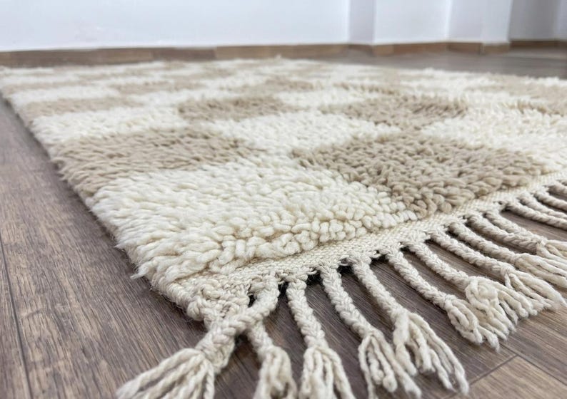 May include: A beige and cream-colored rug with a geometric pattern. The rug has a shaggy texture and a fringe along one edge. The rug is made of a soft material and is suitable for a living room or bedroom.
