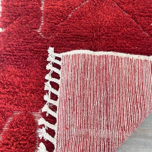 May include: A red, textured rug with a geometric design. The rug has a thick, plush pile and a contrasting white fringe. The underside of the rug is visible, showing a striped pattern in red and white.