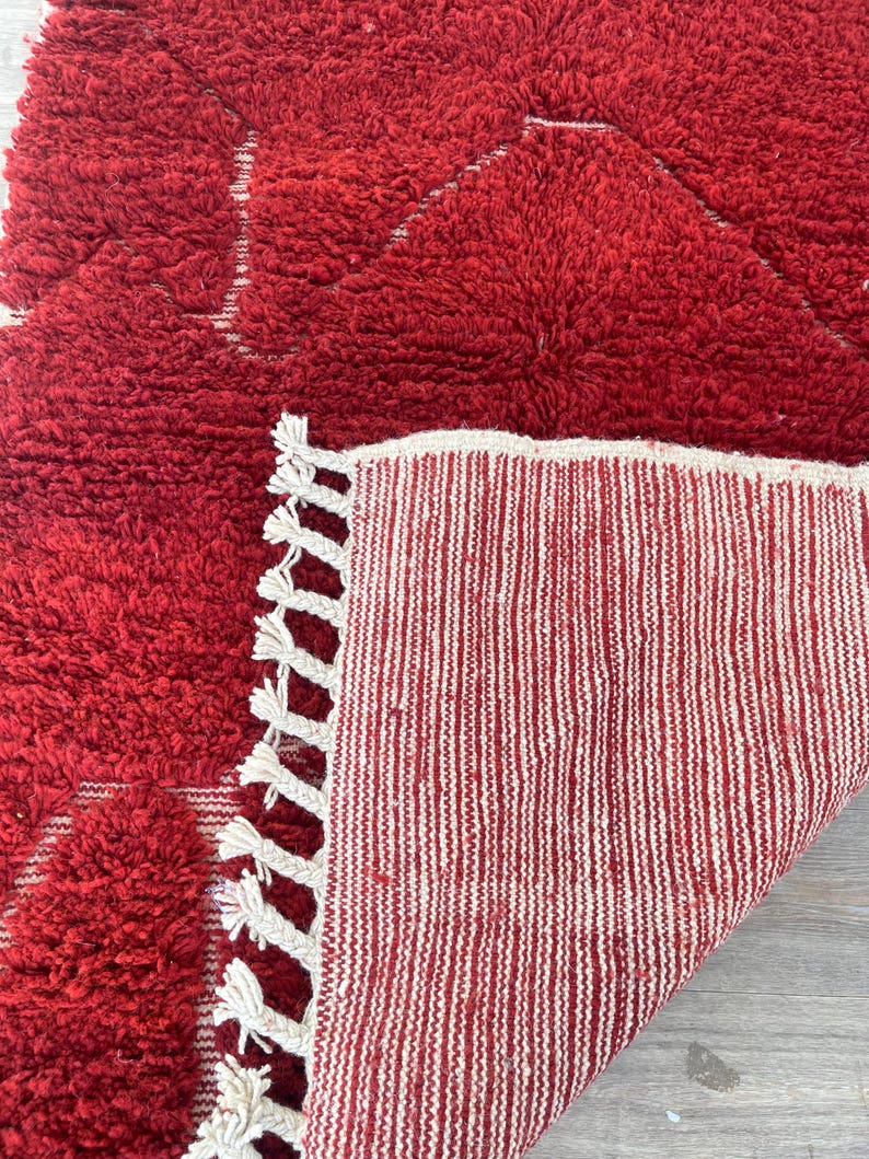 May include: A red, textured rug with a geometric design. The rug has a thick, plush pile and a contrasting white fringe. The underside of the rug is visible, showing a striped pattern in red and white.