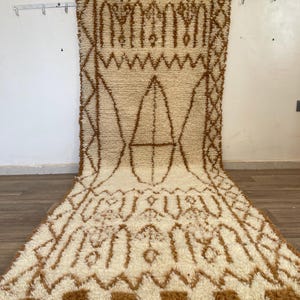 May include: An off-white rectangular rug with a brown geometric pattern. The rug has a thick, textured pile and a fringe along the bottom edge. The design features repeating shapes and lines.