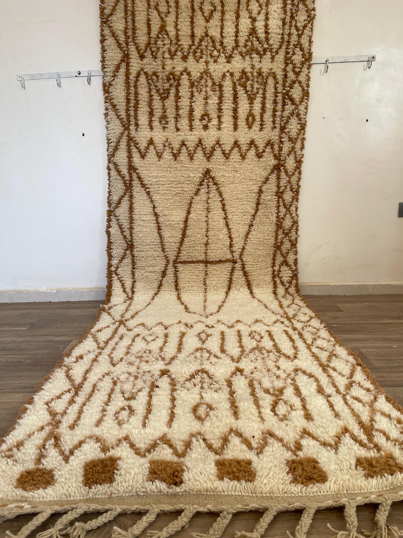 May include: An off-white rectangular rug with a brown geometric pattern. The rug has a thick, textured pile and a fringe along the bottom edge. The design features repeating shapes and lines.