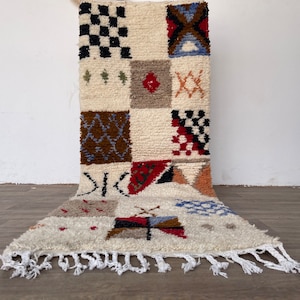 Puede incluir: Una alfombra de lana blanca con un patrón geométrico en tonos de rojo, azul, marrón y negro. La alfombra tiene un borde de flecos.