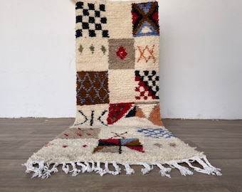 May include: A white wool rug with a geometric pattern in shades of red, blue, brown, and black. The rug has a fringe border.