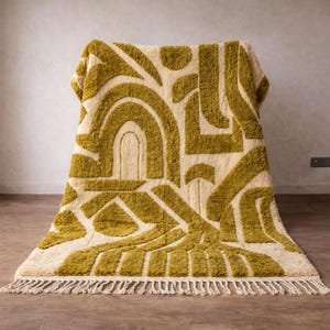 May include: A rectangular rug with a cream-colored background and a geometric pattern in olive green. The design features arches, lines, and shapes. The rug has a fringe along the bottom edge.