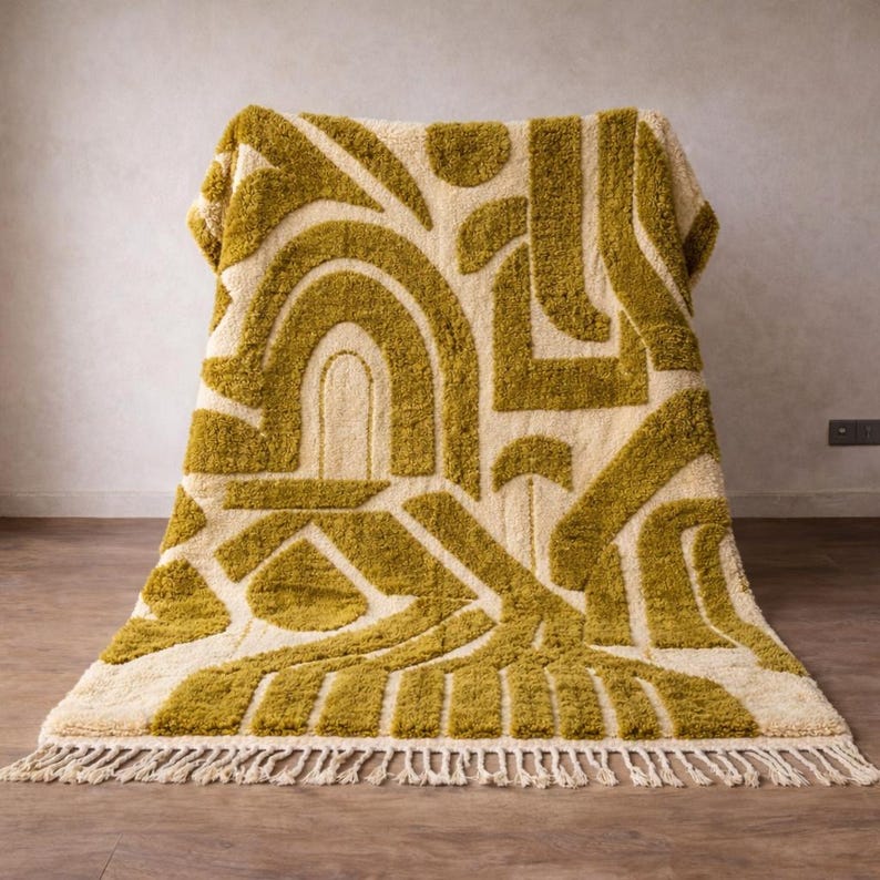 May include: A rectangular rug with a cream-colored background and a geometric pattern in olive green. The design features arches, lines, and shapes. The rug has a fringe along the bottom edge.