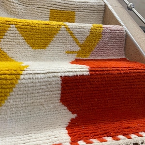 May include: A white, yellow, orange, and pink patterned rug is placed on a set of stairs. The rug is made of a thick, textured material and has a geometric design.