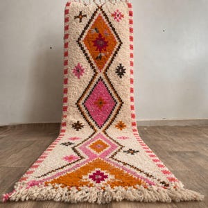 May include: A cream-colored, rectangular rug with a geometric design. The rug features diamond shapes in orange and pink, accented with brown and pink starbursts. The edges are trimmed with a pink border and fringe.