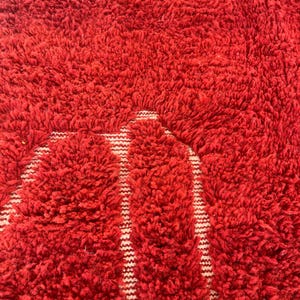 May include: Close-up of a vibrant red, textured rug with a shaggy pile. The rug features a pattern of white vertical stripes against the red background, creating a bold contrast. The thick, plush texture suggests a comfortable and cozy feel.