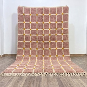 Puede incluir: Una alfombra rectangular con una base de color rosa palo y un patrón de cuadrícula formado por líneas blancas. Pequeños cuadrados de color amarillo mostaza se sitúan en cada intersección de las líneas blancas. La alfombra tiene flecos.