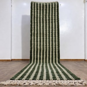 May include: A long, rectangular rug with vertical stripes in olive green and cream. The rug is displayed against a white wall and has a fringed edge. The rug is made of a woven material.