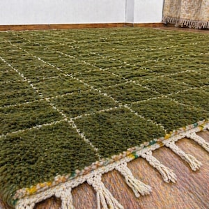 May include: A green, textured rug with a grid pattern of white lines. The rug has a fringe of cream-colored tassels along one edge. The rug is made of a thick, plush material and is suitable for a living room or bedroom.