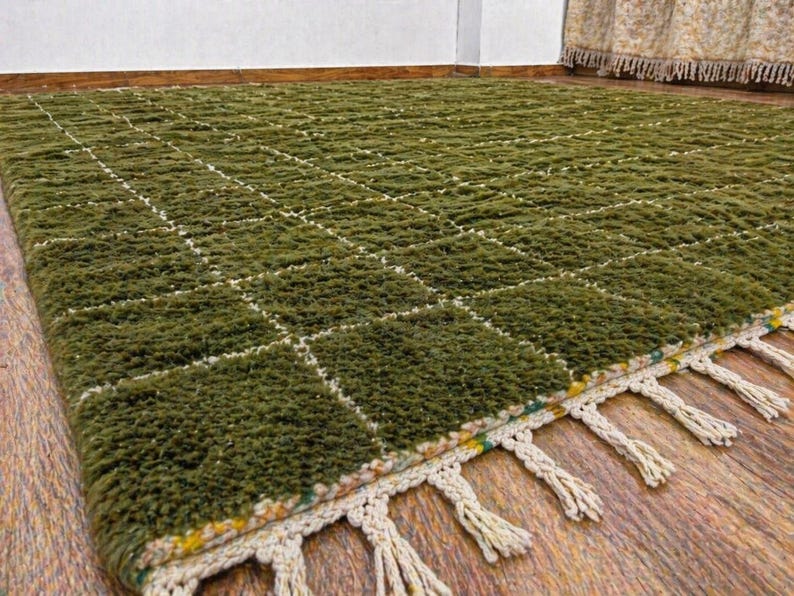 May include: A green, textured rug with a grid pattern of white lines. The rug has a fringe of cream-colored tassels along one edge. The rug is made of a thick, plush material and is suitable for a living room or bedroom.