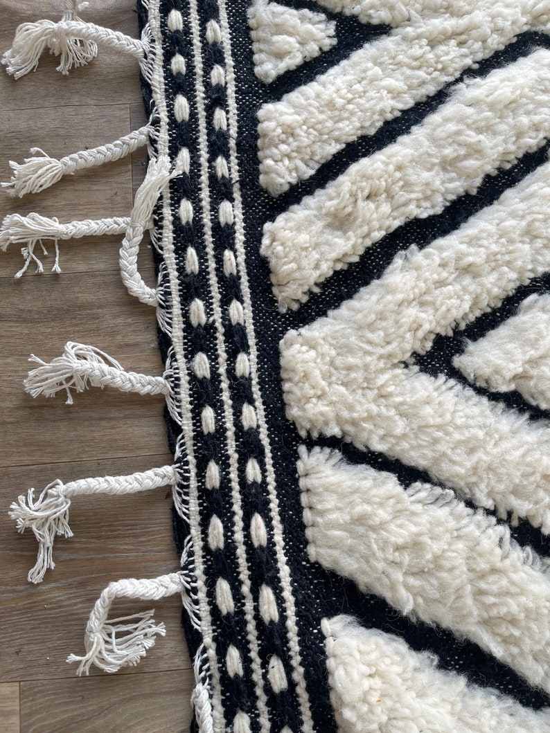 May include: Black and white rug with a geometric pattern and fringe. The rug is made of a thick, fluffy material.
