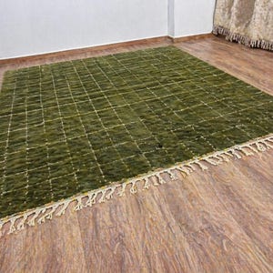 May include: A rectangular olive green rug with a grid pattern and cream-colored fringe. The rug is made of a textured material and is displayed on a wooden floor.
