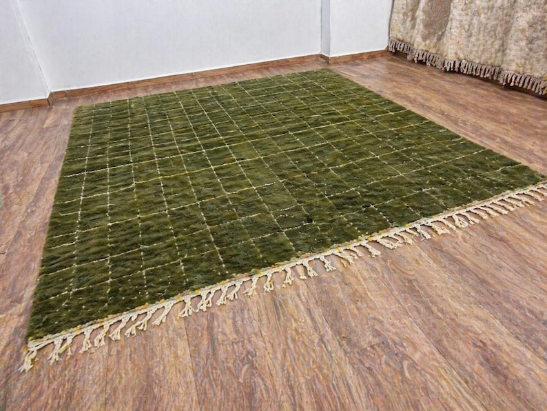 May include: A rectangular olive green rug with a grid pattern and cream-colored fringe. The rug is made of a textured material and is displayed on a wooden floor.