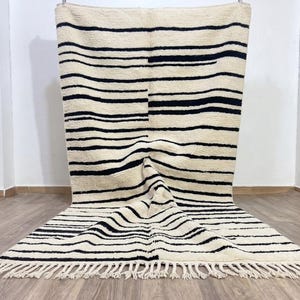 May include: A rectangular area rug featuring a cream-colored base with horizontal black stripes of varying widths. The rug has a thick, textured pile and is finished with a fringe of cream-colored tassels along the bottom edge of the fabric.