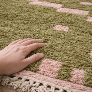 Puede incluir: Primer plano de una alfombra texturizada con un patr&oacute;n de rect&aacute;ngulos y cuadrados rosas sobre un fondo verde. La alfombra tiene una franja a lo largo del borde inferior. Una mano descansa sobre la alfombra.