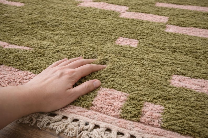 Puede incluir: Primer plano de una alfombra texturizada con un patr&oacute;n de rect&aacute;ngulos y cuadrados rosas sobre un fondo verde. La alfombra tiene una franja a lo largo del borde inferior. Una mano descansa sobre la alfombra.