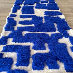 Puede incluir: Una alfombra rectangular, tejida a mano, con un fondo blanco y un patrón geométrico audaz en azul vibrante. El diseño presenta formas irregulares y esponjosas, creando un efecto texturizado y visualmente impactante. La alfombra es probablemente de lana.
