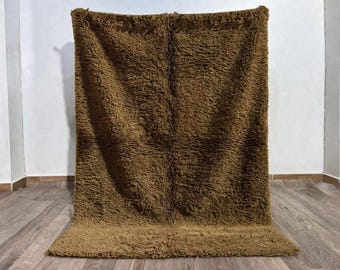 May include: A brown, shaggy rug with a thick, textured pile. The rug is rectangular and hangs against a white wall, with a portion resting on a wooden floor. The color is a warm, earthy tone, suitable for home decor.