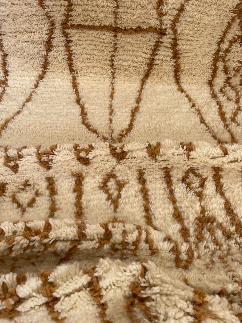 May include: Close-up of a beige and brown patterned rug. The rug features a repeating geometric design with brown lines on a beige background. The texture appears soft and plush, with a high pile.