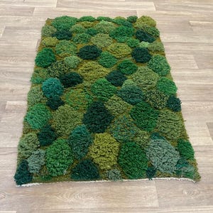 May include: A rectangular rug with a textured, moss-like design in various shades of green. The rug's surface is covered with tufted, rounded shapes, creating a three-dimensional effect. The rug is placed on a light-coloured wooden floor.