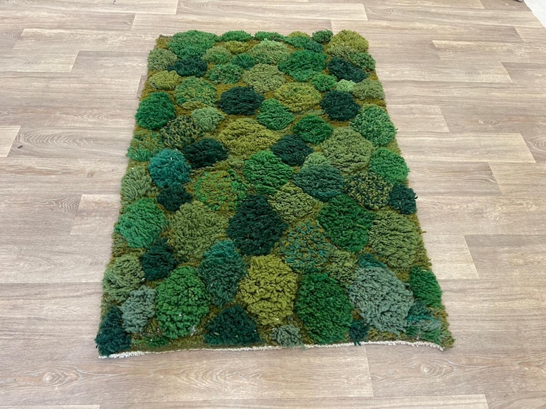 May include: A rectangular rug with a textured, moss-like design in various shades of green. The rug's surface is covered with tufted, rounded shapes, creating a three-dimensional effect. The rug is placed on a light-coloured wooden floor.