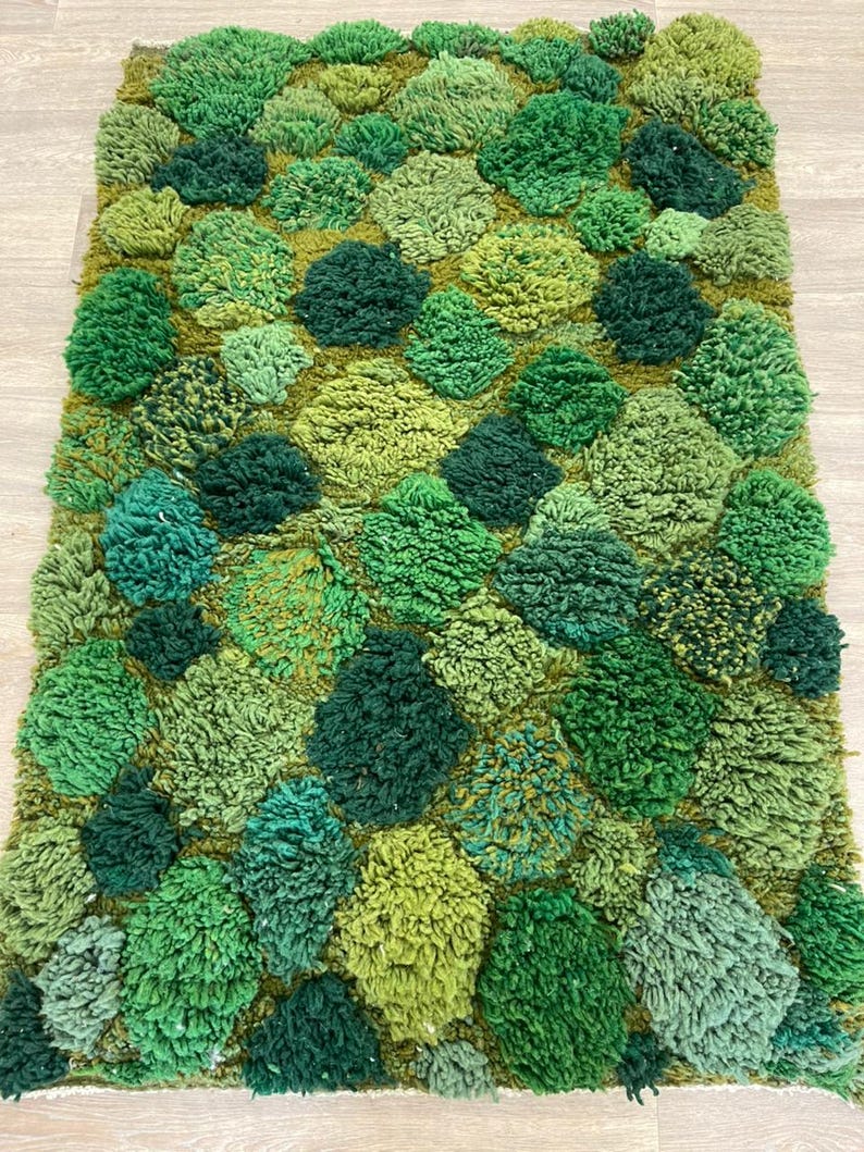 May include: A rectangular rug with a textured, tufted design in various shades of green. The rug features a pattern of raised, rounded shapes, creating a three-dimensional effect. The colour palette ranges from light to dark greens.