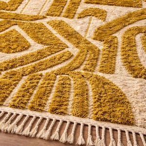 May include: Mustard yellow and cream-colored area rug with a geometric pattern. The rug features a textured, tufted design with abstract shapes and a fringe border. The rug is made of a soft material.