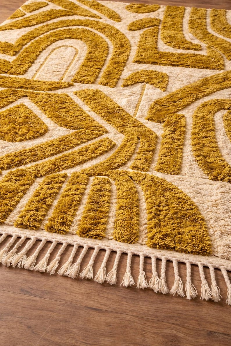 May include: Mustard yellow and cream-colored area rug with a geometric pattern. The rug features a textured, tufted design with abstract shapes and a fringe border. The rug is made of a soft material.