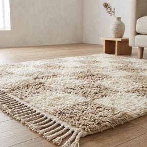 May include: A rectangular, shaggy rug with a geometric pattern of alternating beige and cream stripes. The rug has a fringed edge and is placed on a hardwood floor. The rug is a neutral color and would fit in a living room.