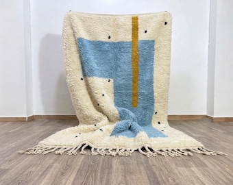 May include: A thick, cream-colored wool rug featuring a large, light blue rectangular block and a vertical mustard yellow stripe. The rug is decorated with small, scattered black dots and finished with long, knotted fringe along the bottom edge.