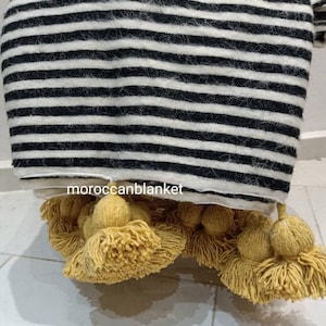 May include: Black and white striped throw blanket with yellow pom pom trim. The blanket is made of wool and has a soft, fuzzy texture. The pom poms are attached to the bottom edge of the blanket.