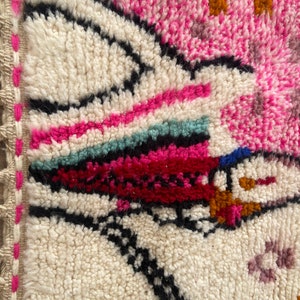 Op de afbeelding: Een kleurrijk Marokkaans tapijt met een witte achtergrond en geometrische patronen in roze, rood, oranje, zwart en blauw.