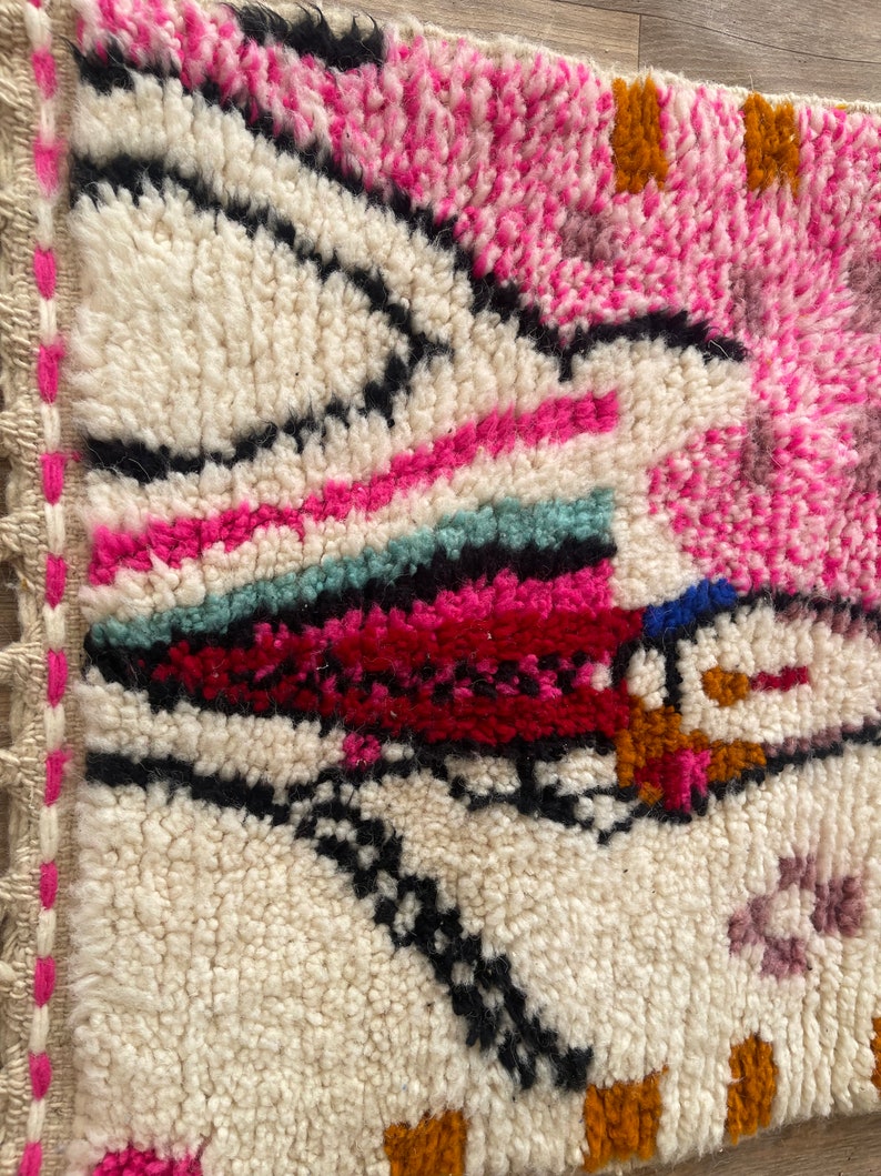 Op de afbeelding: Een kleurrijk Marokkaans tapijt met een witte achtergrond en geometrische patronen in roze, rood, oranje, zwart en blauw.