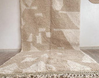May include: A beige and white geometric patterned rug with a textured, plush surface. The rug features a repeating pattern of shapes and a fringe border. The rug is displayed on a light-colored floor.
