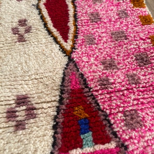 Op de afbeelding: Close-up van een roze en wit geweven tapijt met geometrische patronen in rood, bruin en blauw. Het tapijt heeft een pluche textuur en is gemaakt van wol.