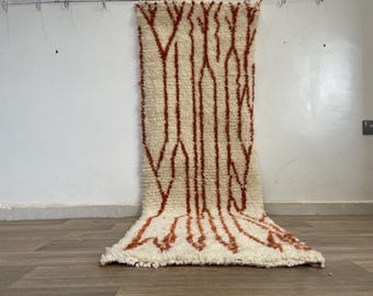 May include: Cream-coloured, rectangular rug with a geometric pattern in shades of rust. The rug has a shaggy texture and is hanging against a white wall. The design features vertical lines and a textured appearance, suitable for home decor.
