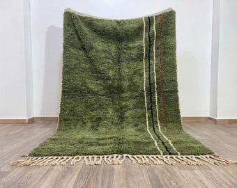 Op de afbeelding: Een groot, rechthoekig, olijfgroen hoogpolig tapijt met een dikke textuur. Het tapijt is aan de rechterkant versierd met verticale strepen in crème, zwart en dunne rode lijnen, afgewerkt met lange, crèmekleurige franjes aan de onderkant.