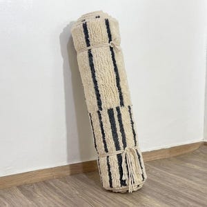 May include: A rolled-up area rug featuring a cream-colored shaggy texture with vertical dark charcoal gray stripes. The rug is secured with two light-colored rope ties, and a fringe of tassels hangs from the bottom edge of the rolled fabric.