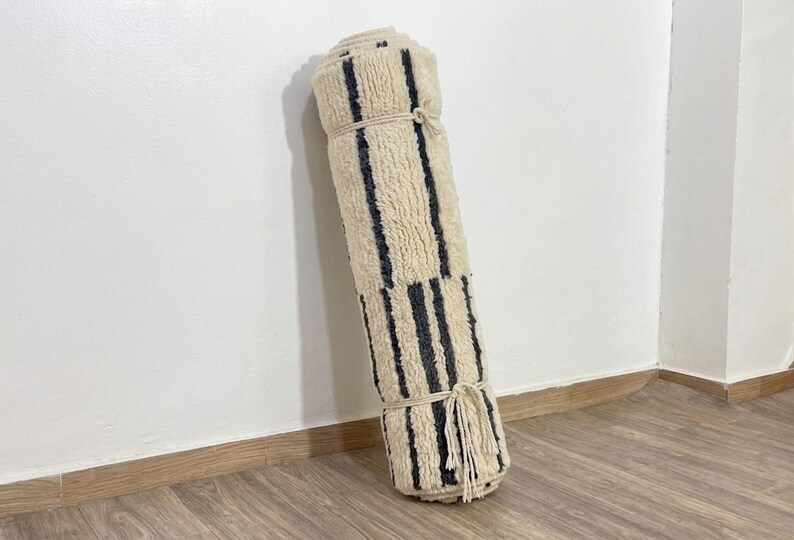 May include: A rolled-up area rug featuring a cream-colored shaggy texture with vertical dark charcoal gray stripes. The rug is secured with two light-colored rope ties, and a fringe of tassels hangs from the bottom edge of the rolled fabric.