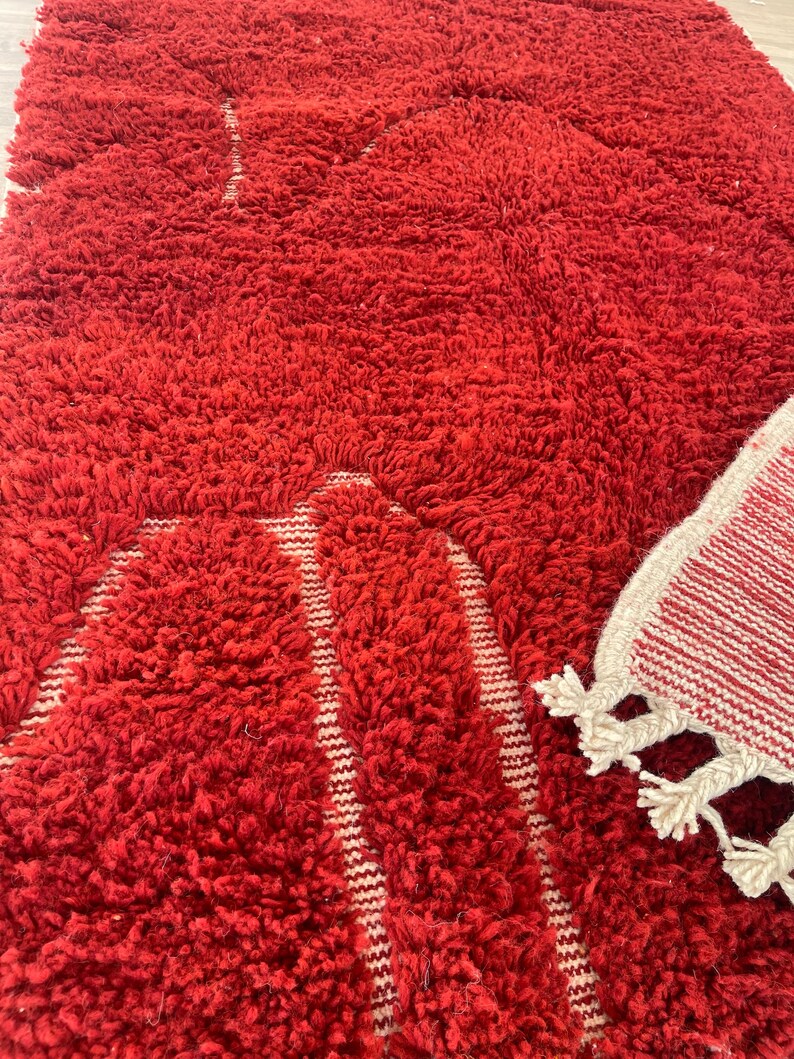 May include: A close-up of a vibrant red, shaggy rug with a textured surface. The rug has a geometric design with white stripes. A smaller rug with white tassels is visible in the corner.