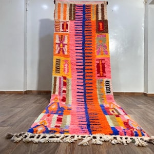 Pode incluir: Um tapete retangular vibrante com um design geométrico colorido. O tapete apresenta uma faixa azul vertical central, rodeada por blocos laranja, rosa e castanhos. A borda inferior tem uma franja branca. O tapete é exibido em um piso de madeira.